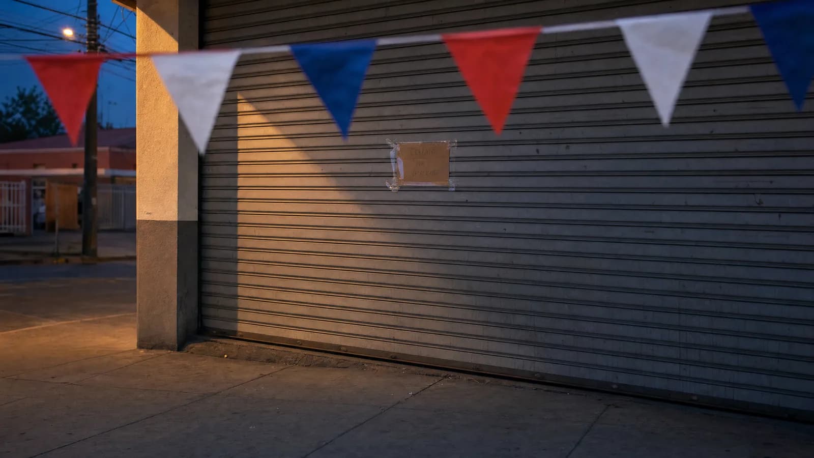 Cortina metálica gris de un local comercial bajada hasta el suelo al anochecer, con un cartel de cartón escrito a mano pegado con cinta a la altura de la vista y guirnaldas de banderines chilenos cruzando la vereda por delante.