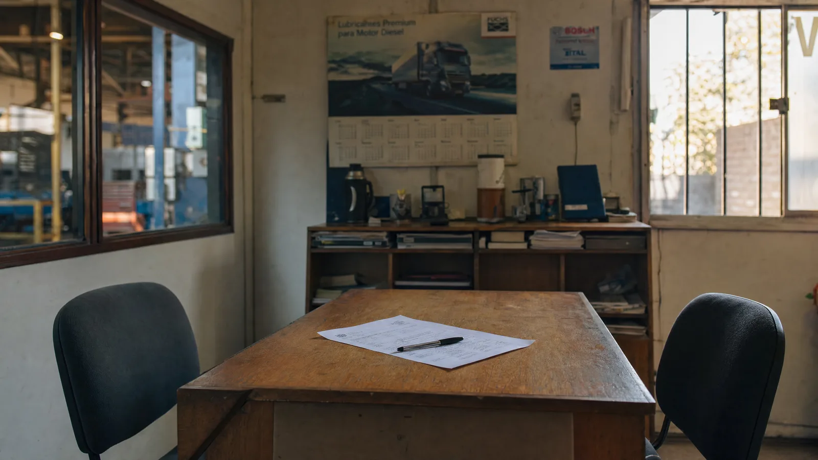 Escritorio de madera vacío en la oficina de un taller, con un documento impreso y un lápiz encima y dos sillas de oficina a cada lado. Al fondo, un mueble con carpetas y un termo, y en la pared un afiche de lubricantes con un calendario.