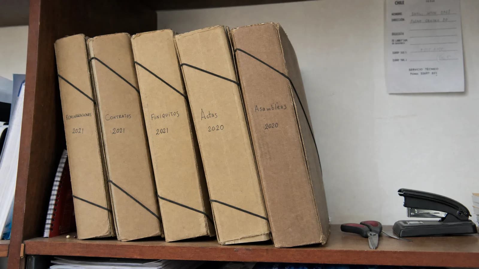 Cinco carpetas de cartón atadas con elástico sobre un estante de madera, rotuladas a mano Remuneraciones 2021, Contratos 2021, Finiquitos 2021, Actas 2020 y Asambleas 2020, con una corchetera y un perforador al lado