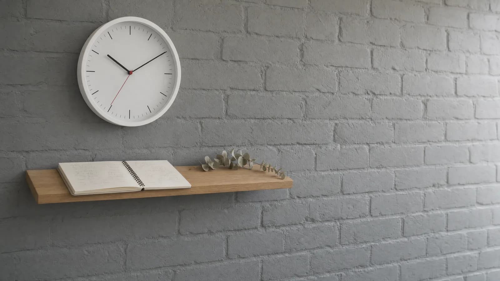 Reloj de pared redondo blanco con segundero rojo, sin números en la esfera, colgado sobre un muro de ladrillo pintado de gris claro, y debajo una repisa angosta de madera con una libreta espiral abierta y una rama seca de eucalipto apoyada al lado.