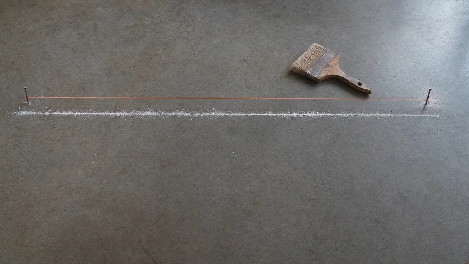 Línea de tiza blanca recién trazada sobre un piso de hormigón pulido gris, con un cordel tensado entre dos clavos a un costado y una brocha de obra apoyada en el suelo.